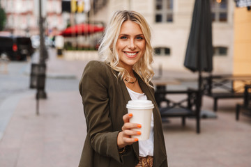 Image of gorgeous woman wearing jacket holding takeaway coffee in paper cup, while walking through city street