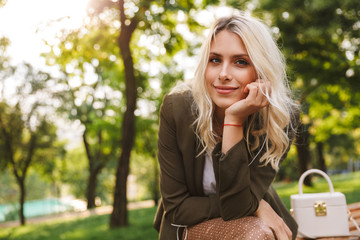 Image of adorable woman 20s smiling at camera, while sitting on bench in park with trees