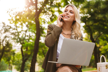 Image of pleased woman 20s using silver laptop and talking on mobile phone, while sitting on bench in park