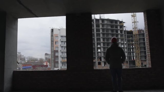 Adult man in orange hemlet is at a construction site, looks out the window of an unfinished house, daylight, sow motion, dolly shot, gimbal