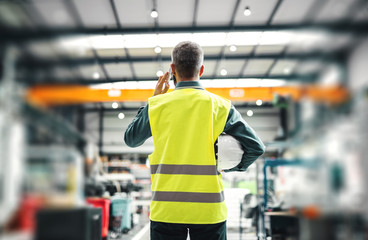 A rear view of an industrial man engineer with smartphone in a factory, working.