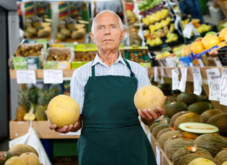 Positive senior male owner of greengrocery shop in apron offering fresh fruits and vegetables