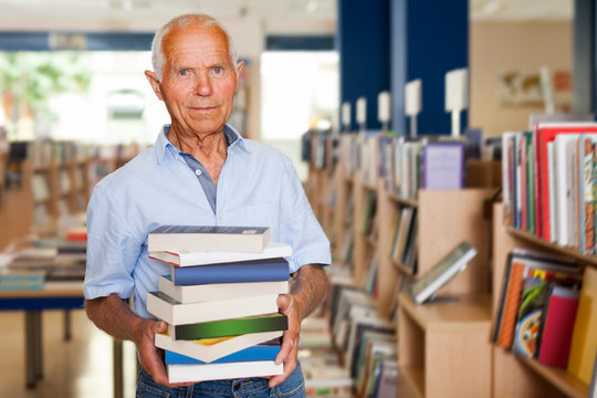 Portrait of interested older man in library with pile of books in hands