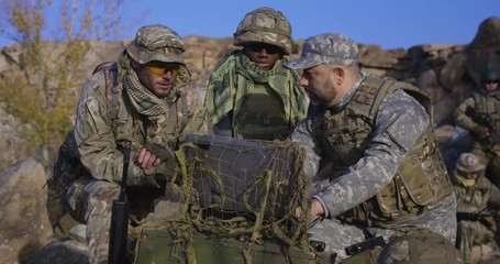 Medium shot of fully equipped and armed african american soldier looking at a computer and another talking on a radio in the field