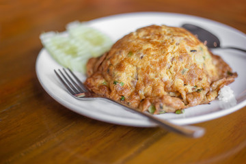 The fried omelette with white pork and green pasley coriender covering white jasmine rice in the white plastic plate with green cucumber on the wooden table in the restaurant.