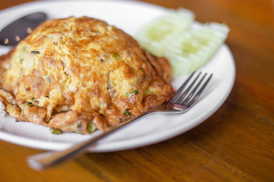 The fried omelette with white pork and green pasley coriender covering white jasmine rice in the white plastic plate with green cucumber on the wooden table in the restaurant.