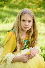 Blond young girl posing in a yellow green dress sitting on the grass with dandelions yellow flowers