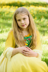 Blond young girl posing in a yellow green dress sitting on the grass with dandelions yellow flowers