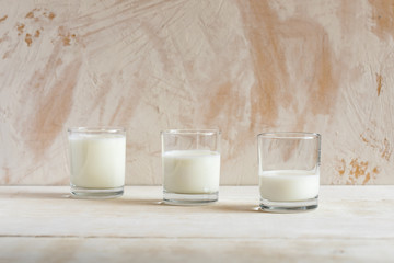 Three glass cups with kefir are standing on white table on white textured background .