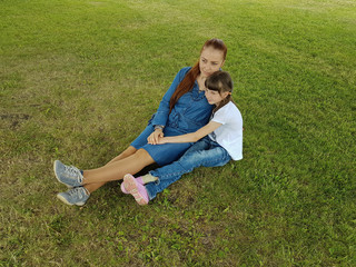 a beautiful young woman in a blue denim dress is sitting with her daughter in the Park under a big tree on a summer day.