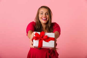 Photo of european chubby woman 20s wearing red dress smiling and holding gift box, isolated over pink background