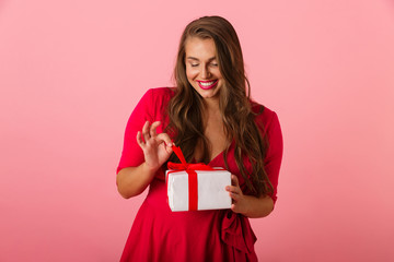 Photo of beautiful chubby woman 20s wearing red dress smiling and holding gift box, isolated over pink background