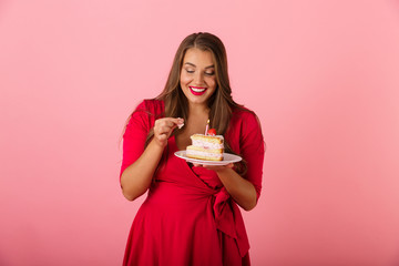 Excited hungry young woman isolated over pink wall background holding cake.