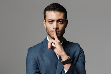 Image of adult arabic businessman 30s in formal suit holding index finger on lips, isolated over gray background