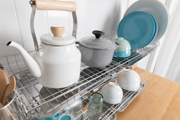 Clean dishes drying on metal dish rack on light background. Kitchen utensils and dishware on wooden shelf. Kitchen interior background.Text space.