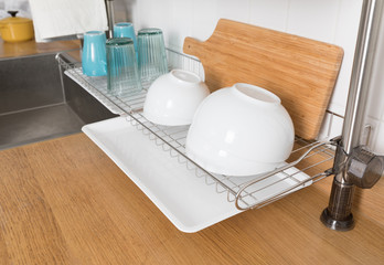 Clean dishes drying on metal dish rack on light background. Kitchen utensils and dishware on wooden shelf. Kitchen interior background.Text space.