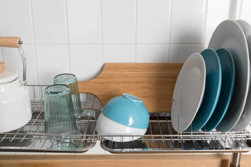 Clean dishes drying on metal dish rack on light background. Kitchen utensils and dishware on wooden shelf. Kitchen interior background.Text space.