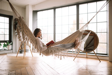 Dark-haired girl dressed in pants, sweater and warm slippers reads a book lying in a hammock in a...
