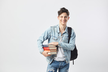 Smiling young dark-haired guy with black backpack  on his shoulder dressed in a white t-shirt, jeans and a denim jacket holds books in his hands on the white background  in the studio