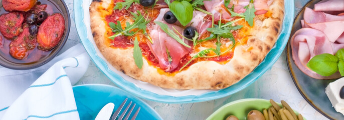 Round large pizza from the oven with prosciutto and olives, arugula and basil. Table setting for lunch or dinner. Table and dishes in blue. Banner.