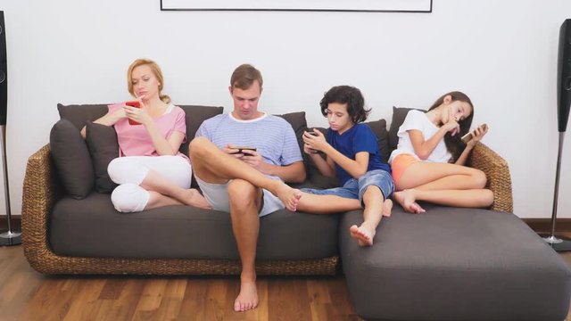 Charming family, mom, dad, daughter and son are watching TV in the living room together, everyone is looking into his phone