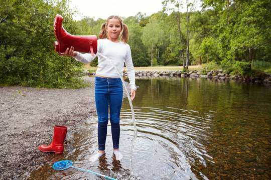 Teenage girl standing in a river wearing socks pours water out of her red wellington boot