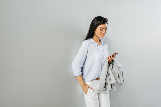 Beautiful elegant Asian woman standing by the white wall and looking at her cell phone.