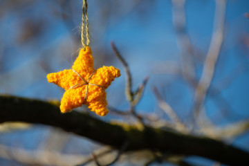 woollen  orange and gold christmas star with twine hanging from tree with blurred branches in the background