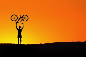 Silhouette of man standing in action lifting bicycle above his head on the meadow with beautiful sunset background,Ride the bike on orange sky scene.Abstract,sport concept.