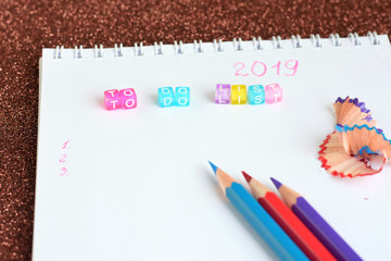 New year 2019 to do list in spiral white paper album. color pencils red blue and violet, colorful plactic letter with selective focus and notebook on shiny sparkle glittery background. back to school.