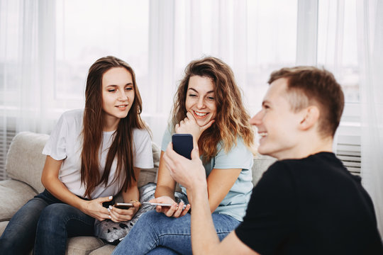 young man showing funny photos or videos on cellphone to his friends, happy people laughing using mobile phone app together sitting in living room