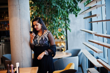 Pretty indian girl in black saree dress posed at restaurant with mobile phone at hand.