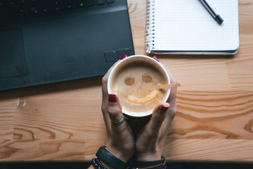 Top view. Student woman holds a hot cup of tea with sea buckthorn Hippophae and a slice of orange. happy smile in a cup. Workplace, notebook with pen and laptop, the working process. coffee shop