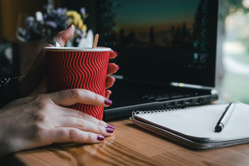 woman holds a hot red cup of tea, coffee. Workplace, notebook with pen and laptop, the working process. coffee shop, space for text, morning work at the computer, study