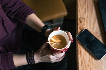 Top view. Student woman holds with two hands a hot cup of tea with sea buckthorn Hippophae and a slice of orange. Workplace, notebook with pen and laptop, mobile phone, smartphone. coffee shop