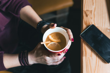 Top view. Student woman holds with two hands a hot cup of tea with sea buckthorn Hippophae and a slice of orange. Workplace, notebook with pen and laptop, mobile phone, smartphone. coffee shop