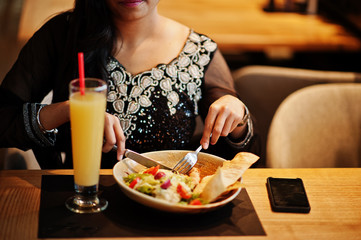 Pretty indian girl in black saree dress posed at restaurant, sitting at table with juice and salad.
