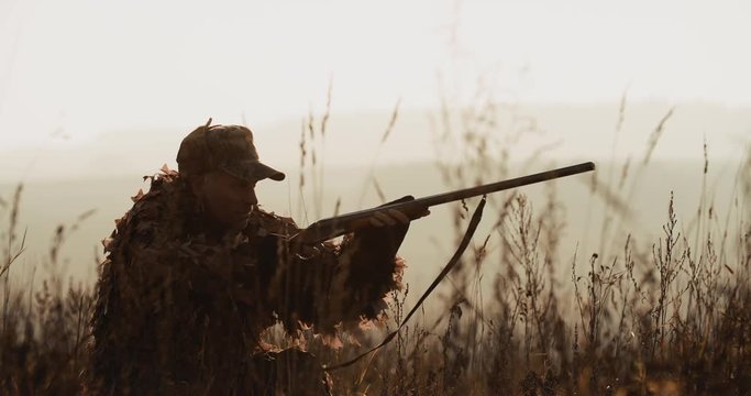 Hunter in hunting equipment lies in wait in the field. Silhouette of man in sunset light find the target and aim with rifle foggy landscape on background