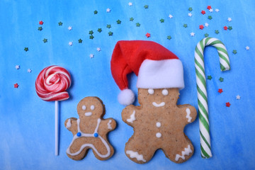 Big and little gingerbread men with lollipops and Santa Claus hat. Confectionery sprinkles around against the blue background. Christmas composition