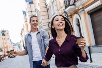 Beautiful woman holds boyfriend by the hand and goes with him along the street of the old town on the background of the sunset. Happy man follows his girlfriend. Follow me concept, female perseverance