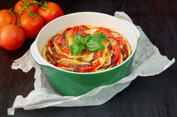 Traditional French dish quiche with eggplants and tomatoes in the baking dish decorated with fresh basil leaves. Homemade vegetable tart on the black background. Side view