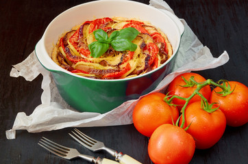 Traditional French dish quiche with eggplants, tomatoes and basil leaves in the baking dish. Homemade vegetable tart on the black wooden background. Side view