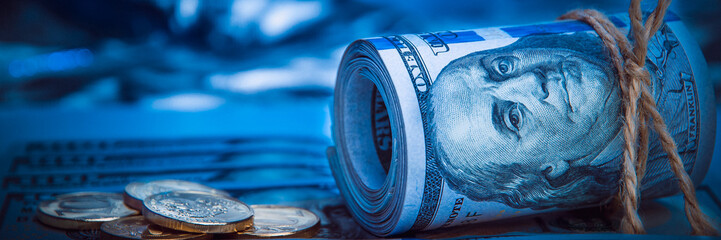 A roll of dollars with coins on the background of scattered one hundred dollar bills in blue light.