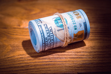 A roll of dollars on a wooden textured table in the dark illuminated by a ray of light.
