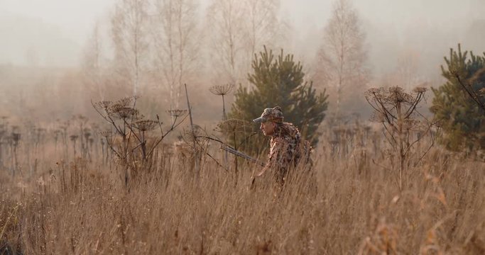 Hunter in hunting equipment with a rifle in his hand sneaks through the dry grass to the target in the field. Sunrise light and foggy landscape