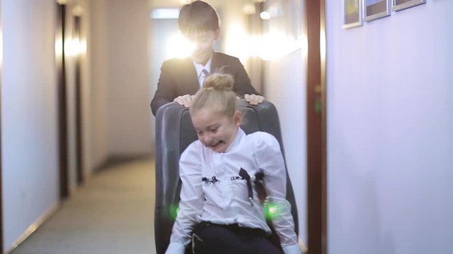 Cute school girl wearing white blouse sitting in office chair. Boy in school uniform driving and laughing. School hallway. Indoors. Children. Fun.
