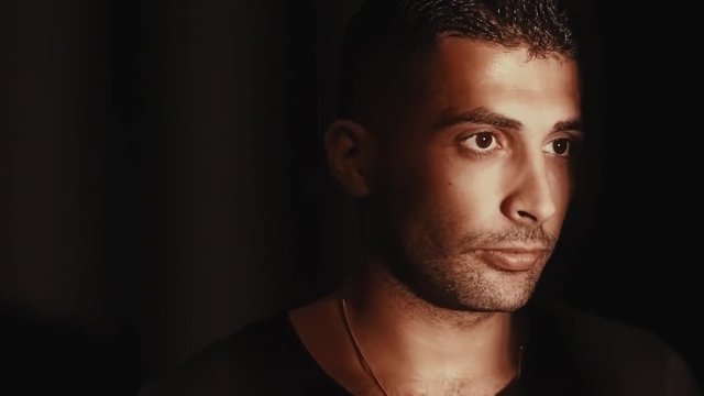 A man of Arab appearance is standing in a dark room, comes to the window, a ray of light falls on his face, he looks into the distance, a tense face
