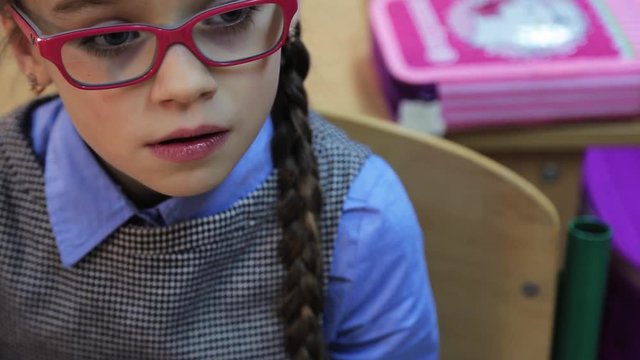 Cute scared little girl with pigtail sitting at desk. Portrait of pretty schoolgirl in red glasses. Close-up. Indoors.