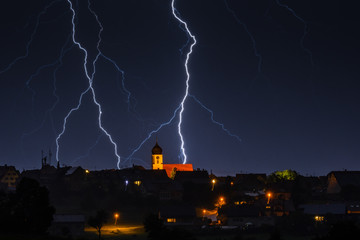 Two lightning strikes near a small village in front of an illuminated church. 