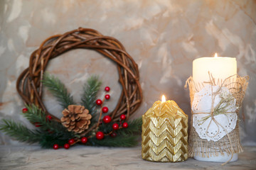 Two Christmas candles, white and gold, stand on a gray background next to a Christmas wreath of branches and trees with red berries and cones. Festive background